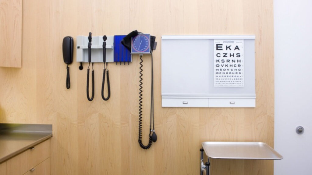 Medical examination room showing a phone, blood pressure cuff and eye examination paper mounted on a light box with a wooden wall in the background