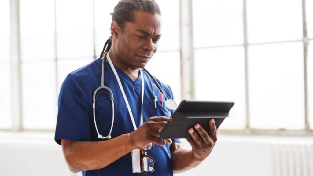 Male healthcare professional looking at a tablet in a hospital hallway with windows
